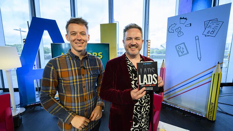 Alan Bissett on the set of Authors Live, smiling and holding his book Lads, Lads, Lads